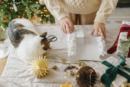 Cute cat helping woman in cozy sweater wrapping christmas gift box on table with modern ornaments on background of decorated stylish christmas tree with lights. Merry Christmas and Happy Holidaysの写真素材
