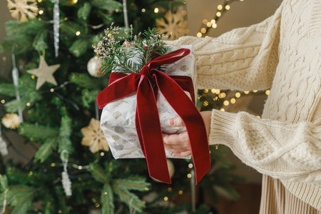 Merry Christmas and Happy Holidays. Hands in sweater holding modern christmas gift box with red ribbon and fir branch on background of stylish christmas tree with lights.の写真素材