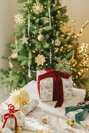 Merry Christmas and Happy Holidays. Stylish christmas gift box with red ribbon, spruce branch and modern ornaments on background of decorated christmas tree. Modern Christmas presentの写真素材
