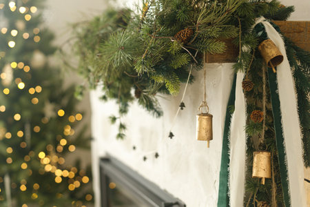 Stylish christmas decorated fireplace mantel with fir branch, bells, ribbon on background of christmas tree with lights in modern living room. Atmospheric holiday at home. Merry Christmas!の写真素材