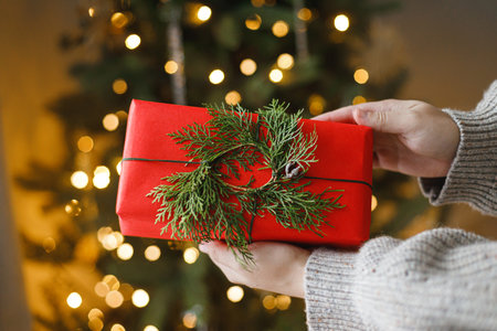 Merry Christmas and Happy holidays! Hands in cozy sweater holding stylish christmas red gift with cedar wreath against golden christmas tree lights. Atmospheric christmas eve, holiday magical timeの写真素材