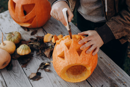 Making a jack o lantern. Hands carving halloween pumpkin on rustic wooden table in autumn garden, hands close up. Fall harvest. Cutting pumpkin. Halloween preparationの写真素材