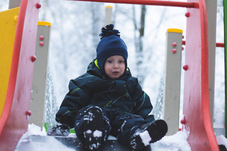 A child rides a slide on the Playground in winter. A boy in a warm coat plays in a snowy winter Park.On the playground, sitting down, rolling down.の写真素材