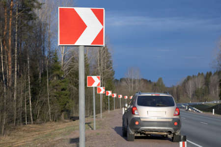 The road sign is a sharp turn of the road to the right and the car in the background stands on the...