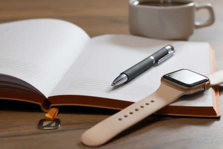 A blank notebook with a pen stands on a wooden office table with a smart watch and a cup of coffee. Copy space.の写真素材