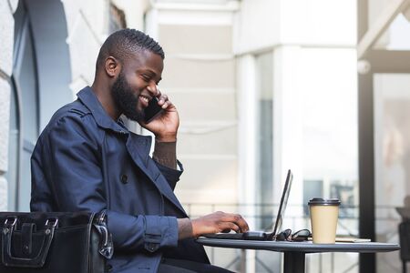 Millennial entrepreneur using laptop while speaking with customers on phone in outdoor restaurant.の写真素材