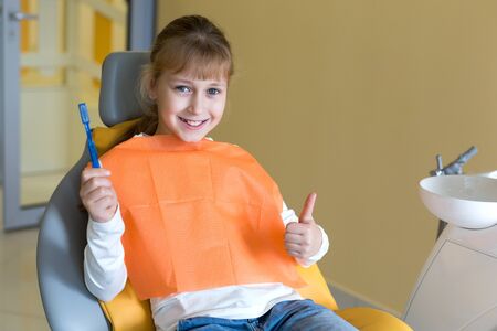 Little cute girl holding a toothbrush and showing a thumb while sitting on a dentist chairの写真素材