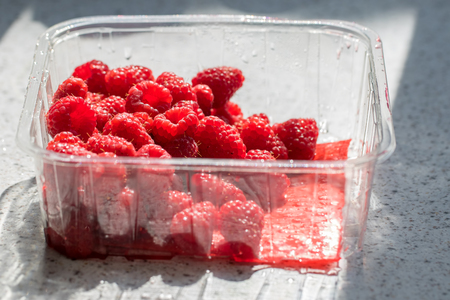 Raspberries in plastic supermarket tub packaging. Healthy ripe red organic fruit in shop bought recyclable container on kitchen worktop.の写真素材