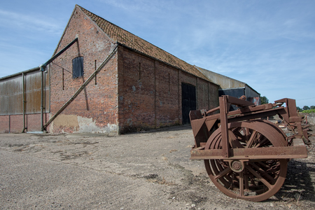 Agricultural shed building and vintage farm machinery in rural Norfolk, UK. Old farming industry land with brick barn and rusty equipment.の写真素材