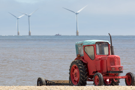 The future of energy production and environmental conservation represented by quirky red diesel beach tractor with offshore clean energy and sustainable resource wind farm turbines in the distance.の写真素材