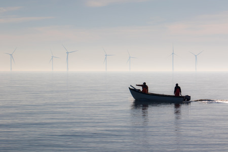 Sustainable resources alternative lifestyle. Small fishing boat with offshore wind turbines. Calm misty morning. Two fishermen heading out to sea. Tranquil landscape scene with beautiful background.の写真素材