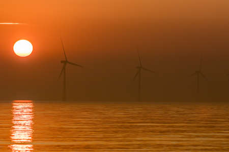 Solar energy and wind power. Offshore windfarm turbines at sunrise. Authentic photographic image of beautiful sun rising over a calm sea on a misty morning. Renewable energy climate change backgroundの写真素材