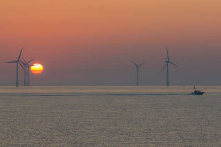 Spiritual landscape. Offshore wind turbines. Peaceful orange sky sunrise scene with small boat crossing the horizon. Clean green energy silhouette with hazy pastel background Tranquil seascape at dawnの写真素材