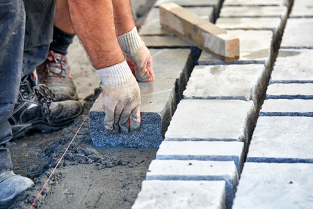 A workman's gloved hands use a hammer to place stone pavers. Worker creating pavement using...