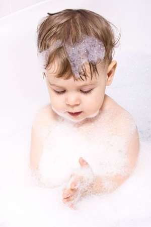 Baby girl sitting in the bath, curious about shampoo foam in her hands.の写真素材