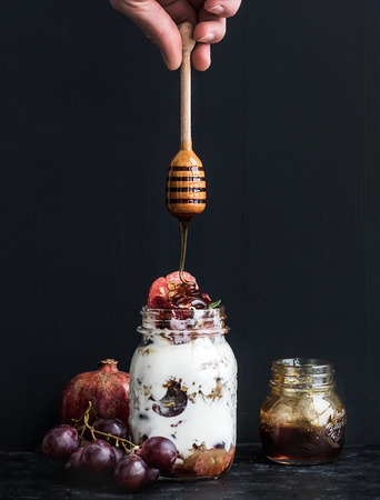 Yogurt and oat granola with grapes, pomegranate and grapefruit in a tall glass jar on black backdrop. Hand pours honey over the jar with a wooden stickの写真素材