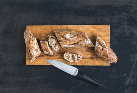 French baguette cut into pieces on a rustic wooden board with knife over dark old painted background. Top viewの写真素材