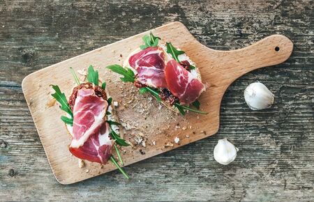 Brushetta set with smoked meat, arugula, garlic and dried tomatoes on a rustic wooden board over a rough wood background. Top viewの写真素材