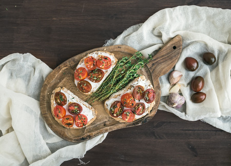 Sandwiches (brushtta) with roasted cherry tomatoes, soft cheese, garlic and herbs on a rustic wooden board and white kitchen towel over a dark wood background. Top viewの写真素材