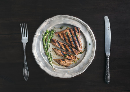 Beef steak cut into pieces with rosemary, knife and fork on a vintage metal plate over a dark wooden backgroundの写真素材