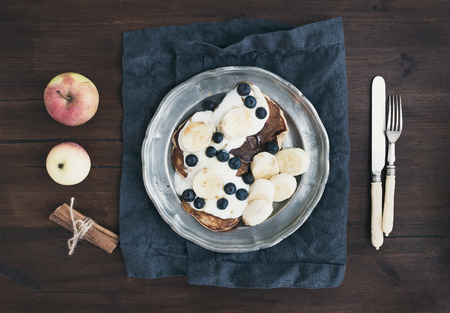 Breakfast set on dark wooden desk: apple and cinnamon pancakes with yogurt, banana, blueberry and maple syrup in a rustic metal plate. Top viewの写真素材
