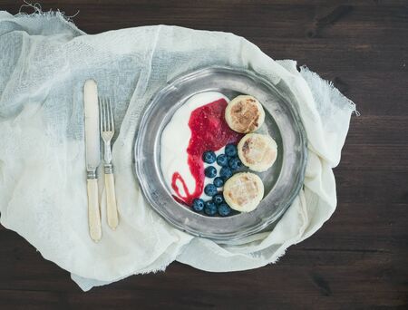 Breakfast set: curd pancakes with yogurt, fresh blueberry and raspberry jam on a vintage metal plate on a piece of white linen fabric over a dark wood background. Top viewの写真素材