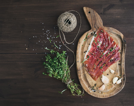 Cured pork meat or prosciutto on a rustic woodem board with garlic, spices and thyme over a dark wood background with a copy space. Top viewの写真素材