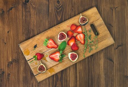 Fresh strawberries, figs, mint leaves, thyme and honey drops on a wooden cutting board over a rough medium on dark wood backgroundの写真素材