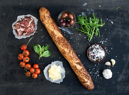 Ingredients for sandwich with smoked meat, baguette, basil, arugula, olives, cherry-tomatoes, parmesan cheese, garlic and spices over black grunge background. Top viewの写真素材