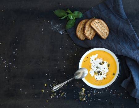 Pumpkin soup with cream, seeds, bread and fresh basil in rustic metal plate on grunge black background. Top view, copy space