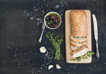 Italian ciabatta bread cut in slices on wooden chopping board with herbs, garlic and olives over dark grunge backdrop, copy space, top viewの写真素材