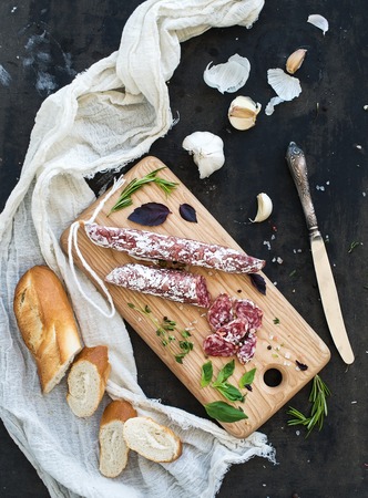 Meat gourmet snack. Salami, garlic, baguette and herbs on rustic wooden board over dark grunge backdrop, top viewの写真素材