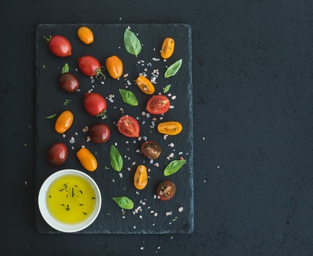 Cherry tomatoes of various color, basil leaves, spices and olive oil on black slate tray over dark grunge background. Top view, copy spaceの写真素材