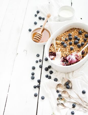 Healthy breakfast. Oat granola berry crumble with fresh blueberries, yogurt and honey in ceramic cooking dish over white rustic backdrop, top viewの写真素材