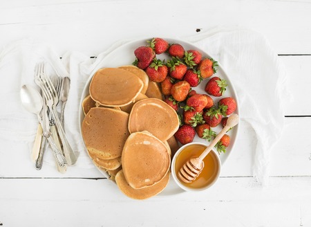 Breakfast plate. Homemade pancakes with fresh strawberry and honey, kitchen napkin, vintage silverware on rustic white background. Top view.の写真素材