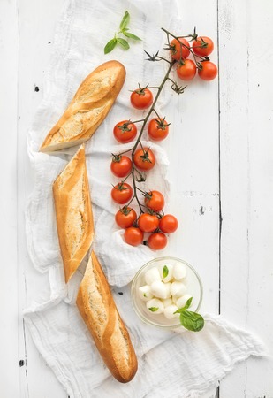 Baguette with banch of cherry-tomatoes, basil and bowl of mozzarella cheese on rustic white wooden backdrop, top view, copy spaceの写真素材
