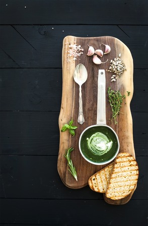 Homemade green spinach cream-soup in a metal scoop with grilled bread slices, herbs, spices on rustic wooden  serving board over black background, top viewの写真素材