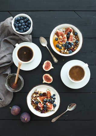 Healthy breakfast set. Bowls of oat granola with yogurt, fresh blueberries and figs, coffee, honey, over black wooden backdrop. Top viewの写真素材