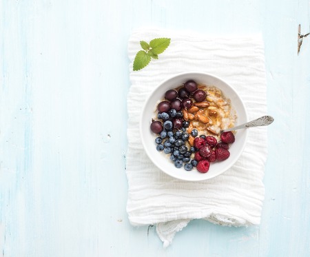 Healthy breakfast set. Bowl of oat porridge with fresh berries, almond and honey over white napkin. Top view, light blue backdrop, copy spaceの写真素材