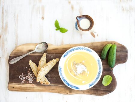 Pumpkin soup with cream, fresh basil, cucumbers and bread in vintage ceramic plate on wooden board over white background, top view, copy spaceの写真素材