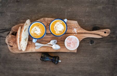 Homemade pumpkin cream soup in enamel mugs with herbs and fresh bread slices on olive serving board over rustic wooden background, top viewの写真素材