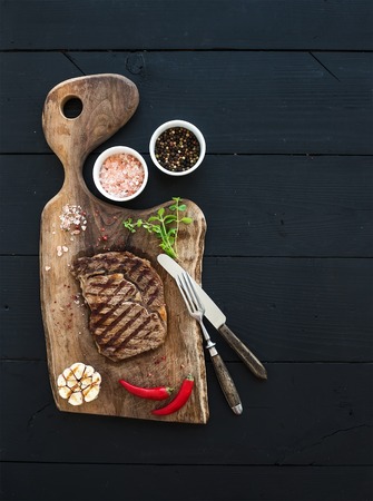 Grilled ribeye beef steak with herbs and spices on walnut cutting board over black wooden background, top view, copy spaceの写真素材