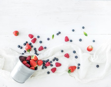 Berry frame with copy space on right. Metal bucket, strawberries, raspberries, blueberries and mint leaves, white wooden background, top view, copy spaceの写真素材
