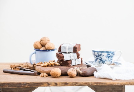 Chocolate brownie slices wrapped in paper and tired with rope, porcelain tea cup and walnuts in enamel mug on rustic wooden table, white background, selective focusの写真素材