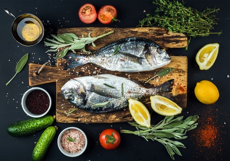 Fresh uncooked dorado or sea bream fish with lemon, herbs, oil, vegetables and spices on rustic wooden board over black backdrop, top viewの写真素材