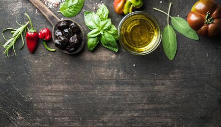 Food background with vegetables, herbs and condiment. Greek black olives, fresh basil, sage, rosemary, tomato, peppers, oil on dark rustic wooden background.  Top view, copy spaceの写真素材