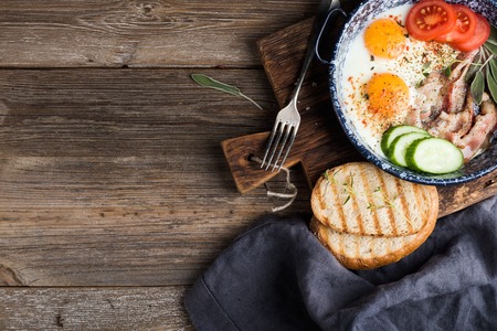 Breakfast set. Pan of fried eggs with bacon, fresh tomato, cucumber, sage and bread on rustic serving board over wooden background, top view, copy spaceの写真素材