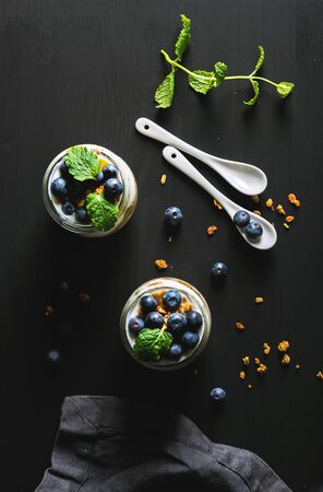 Healthy breakfast set. Yogurt oat granola with blueberries and mint leaves in glass jars on black backdrop, top view, vertical