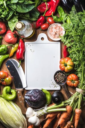 Fresh raw vegetable ingredients for healthy cooking or salad making with white ceramic cutting baoard in center, top view, copy space. Olive oil, spices and knife. Diet or vegetarian food conceptの写真素材