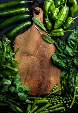 Fresh raw green vegetable ingredients for healthy cooking or salad making with dark wooden cutting baoard in center, top view, copy space. Diet or vegetarian food concept, horizontal compositionの写真素材